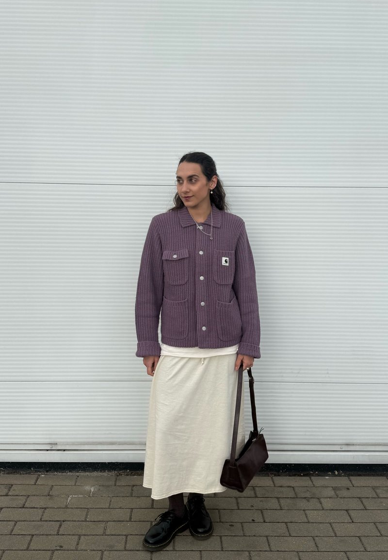 Persona in piedi contro un muro ondulato chiaro, indossa una giacca di maglia viola, gonna crema, scarpe nere e tiene una borsa a tracolla marrone scuro.