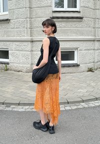 Svart ärmlös topp, orange spetskjol med ojämn fåll, svart slouchy väska och svarta sneakers på en kullerstensgata vid en texturerad vägg.