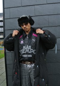 Piumino nero con ricami colorati, camicia a fantasia con il testo "MAS TIEMPO", jeans argentati e cappello di pelliccia. Gli occhiali da sole completano il look.