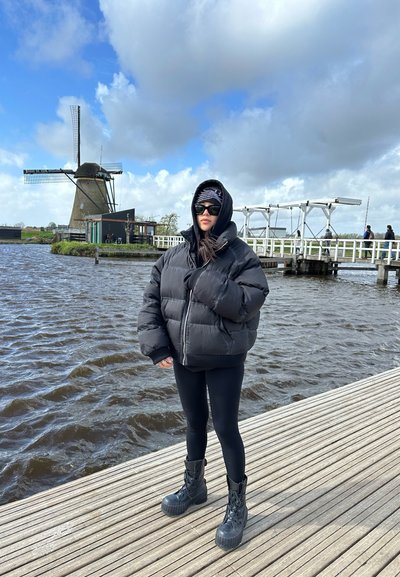 Chaqueta acolchada negra con capucha, leggings negros y botas de combate negras. El fondo presenta un molino de viento y un muelle de madera. Cielos nublados.