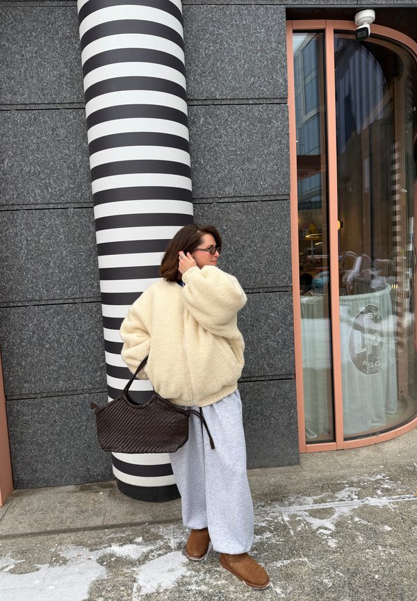 Donna con giacca beige oversize, pantaloni grigi e stivali marroni in piedi accanto a una colonna a strisce bianco e nero fuori da un edificio con grandi finestre.