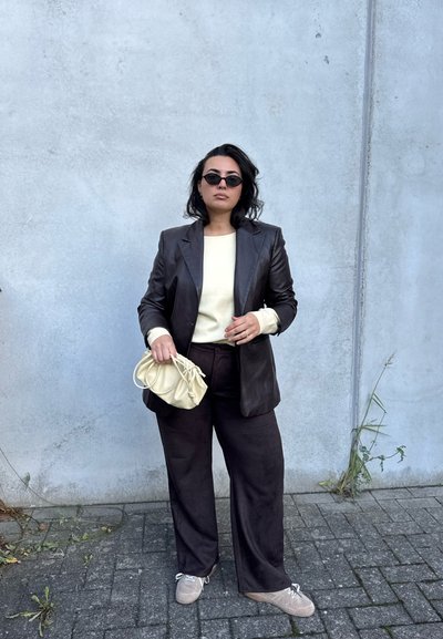 Blazer de cuero marrón sobre un suéter crema claro, pantalones amplios oscuros, sosteniendo un bolso amarillo pálido y usando gafas de sol negras.