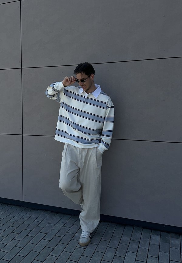 Młody mężczyzna w paskowanej koszuli z długim rękawem, jasnych szerokich spodniach, trampkach i okularach przeciwsłonecznych, opierający się o szarą, kafelkową ścianę z jedną nogą uniesioną.