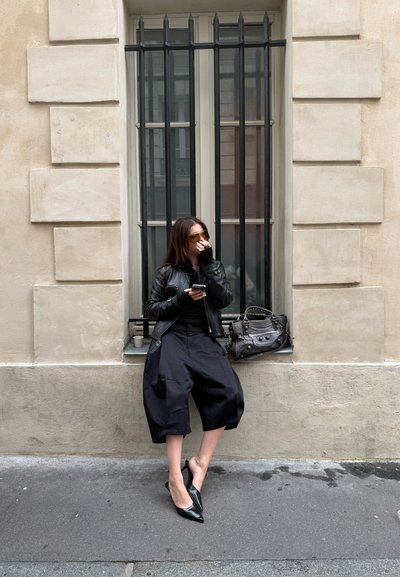 Chaqueta de cuero negra, camisa negra, pantalones cortos negros de gran tamaño y tacones puntiagudos negros. Bolso marrón y gafas de sol amarillas. Sentada junto a una ventana.