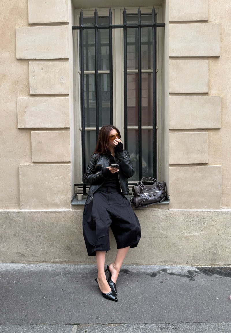 Giubbotto di pelle nera sopra un top nero, pantaloni cropped larghi neri e décolleté lucidi neri a punta. Gli accessori includono occhiali da sole con tonalità dorate e una borsa grigia.