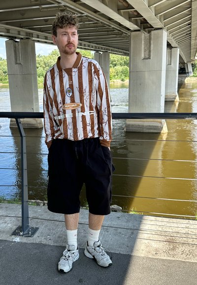 Camisa de manga larga con rayas marrones y blancas, logo en el pecho. Pantalones cortos negros y zapatillas grises claras. Ambiente al aire libre cerca de un río bajo un puente.