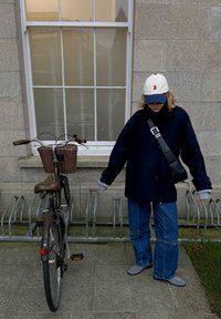Une personne portant une veste bleu marine, un jean et une casquette blanche se tient à côté d'un vélo noir dans un support à vélos métallique devant un bâtiment en pierre.