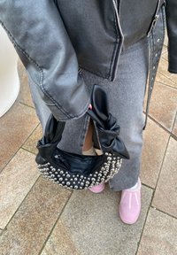 Svart håndveske med knyttet håndtak, prydet med sølvnagler, med en glatt tekstur og rund form. Båret med grå denim.