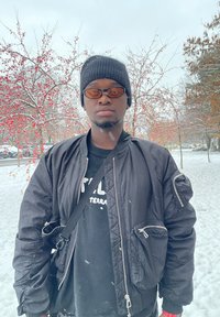 Melna bumbvedera jaka ar rāvējslēdzējiem, valkāta virs melnas T-krekla. Aksesuāri ietver adītu cepuri un tonētas saulesbrilles. Pārsnigta ainava ar sarkaniem ogām.