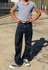 Grå kortærmet henley-shirt, mørkeblå løst siddende jeans, sort bælte og sort-hvide sneakers. Stående pose på fortov.