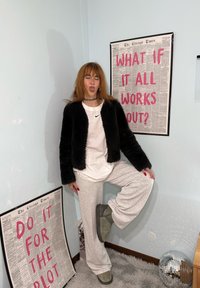 Jeune femme en vêtements décontractés tirant la langue, debout sur une jambe près de deux affiches motivantes encadrées dans un coin aux murs bleu clair.