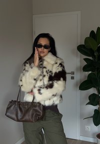 Faux fur-jakke med en blanding av krem og brune mønstre, glidelås og vide ermer. Bærer en mørk brun håndveske. Olivengrønne bukser.