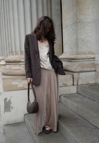 Dolga svetlo rjava satenasta krilo v kombinaciji s temno rjavim suknjičem. Model drži temno rjavo torbico in stoji na sivih kamnitih stopnicah.