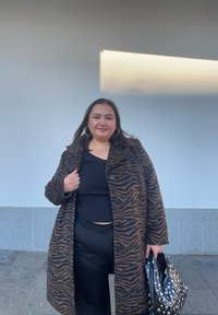 Długi płaszcz w brązowo-czarne prążki zebry z guzikami, noszony na czarnym, prążkowanym topie i spodniach. Całość dopełnia czarna torebka z ćwiekami.