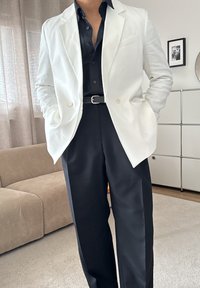 Blazer de lino blanco sobre una camisa negra, combinado con pantalones negros. Los detalles destacados incluyen un cinturón y un bolsillo en el blazer.