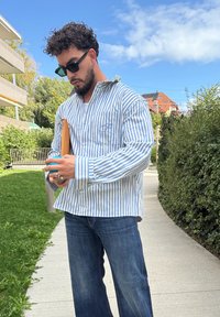 Homem com cabelo encaracolado e barba, vestindo óculos de sol, camisa listrada azul e branca e jeans, segurando um telefone e uma carteira castanha, no exterior, num caminho ensolarado.