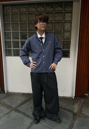 Persona joven con el pelo corto y gafas, lleva una camisa azul sobre una camisa blanca con cuello y corbata, pantalones negros de pierna ancha, de pie con las manos en las caderas.