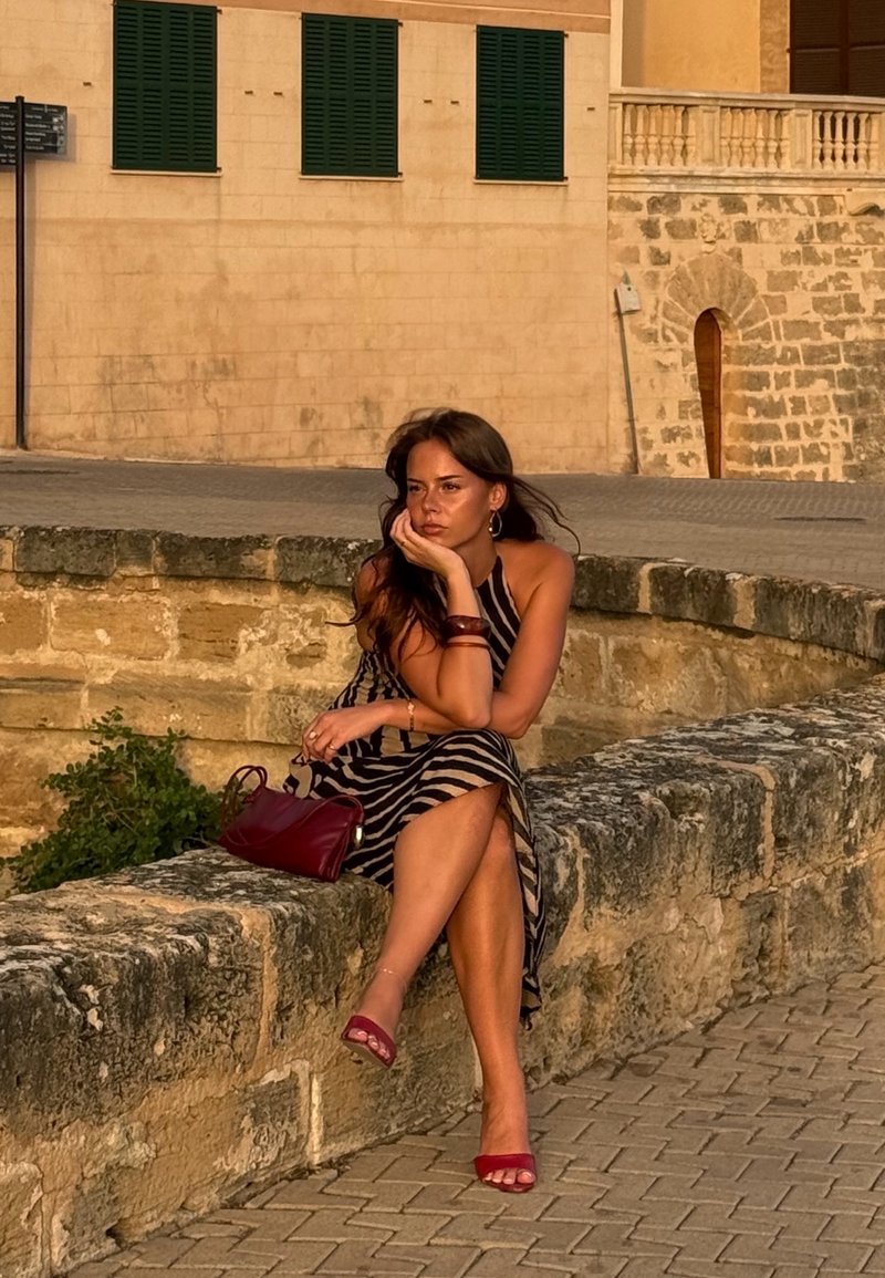 Femme en robe rayée et sandales rouges assise sur un mur de pierre, reposant sa tête sur sa main, un sac rouge à côté d'elle en plein air.
