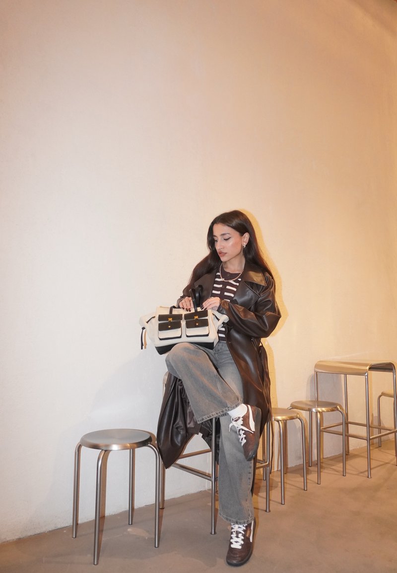 Schwarzer Ledermantel, gestreiftes Oberteil, graue Jeans, hält eine beige Tasche mit schwarzen Akzenten. Grauer Hintergrund und Metallhocker vorhanden.