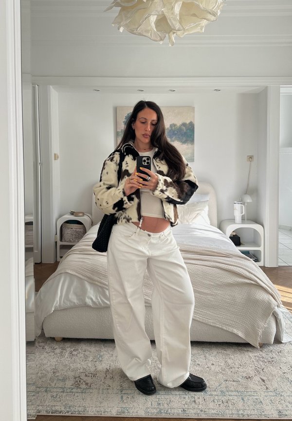 Pitkätummatukkainen nainen, joka pukeutunut mustavalkoiseen kuviolliseen jakkuun, valkoisiin housuihin ja mustiin kenkiin, seisoo makuuhuoneessa ottaen peilistä selfien.