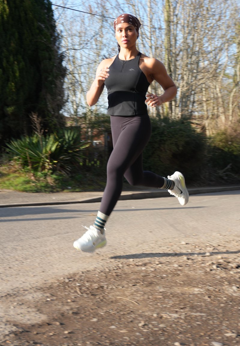 Femme portant une tenue de sport noire et un bandeau, courant en plein air sur une route pavée avec des arbres et des buissons à proximité.