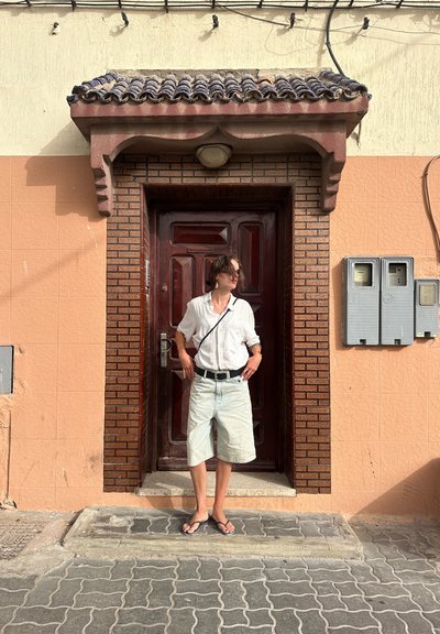 Puerta de madera con acabado rojo oscuro, pared de ladrillo texturizado color beige, azulejos rosa pálido y buzones. Persona con camisa blanca y pantalones cortos de mezclilla se encuentra cerca.