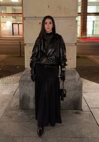 Zwarte leren cropped jas, nauwsluitende zwarte jurk en lange zwarte rok. Handschoenen en een kleine zwarte handtas. Avondsfeer met een stedelijke achtergrond.