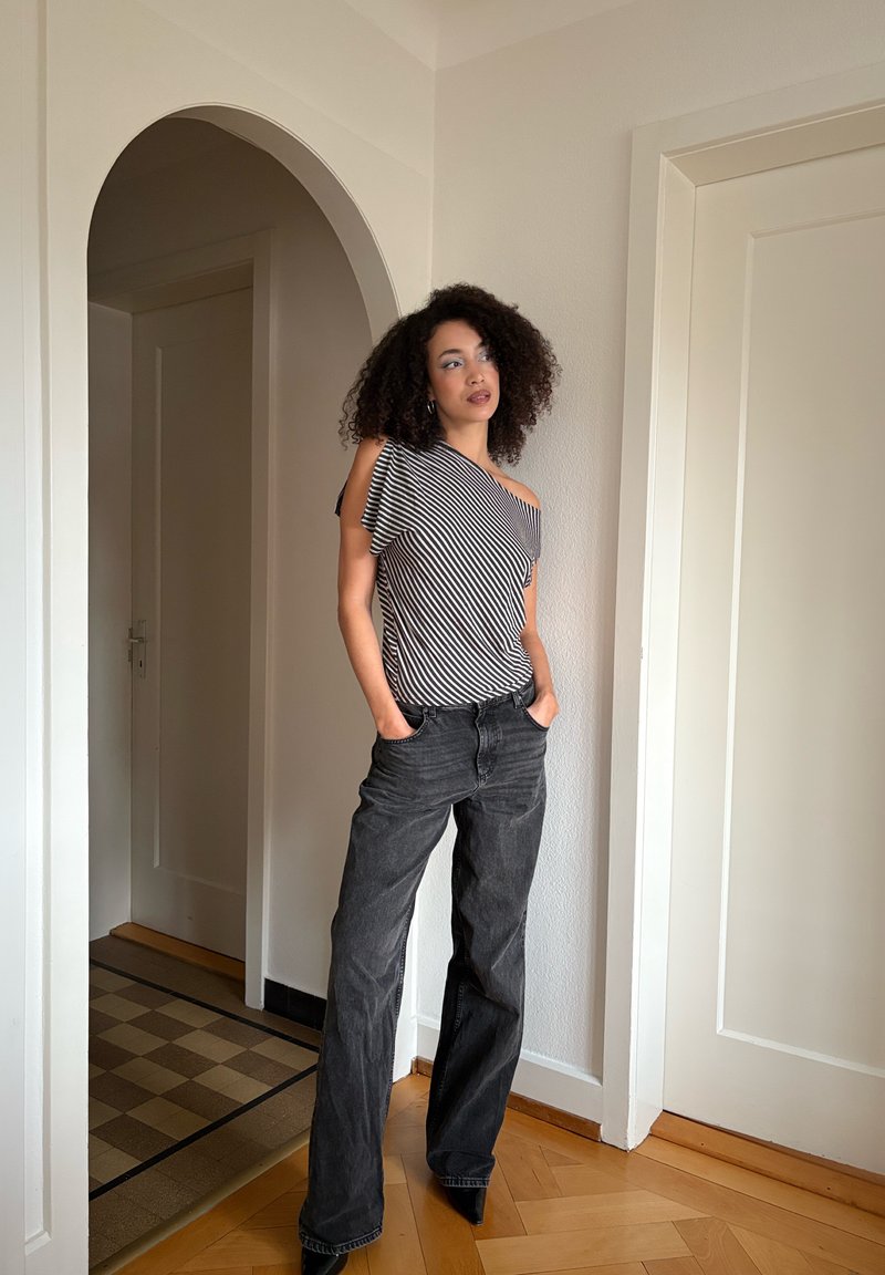 Haut rayé, avec épaules dénudées, en noir et blanc, associé à un jean noir ample taille haute et des escarpins noirs pointus. Pose debout.