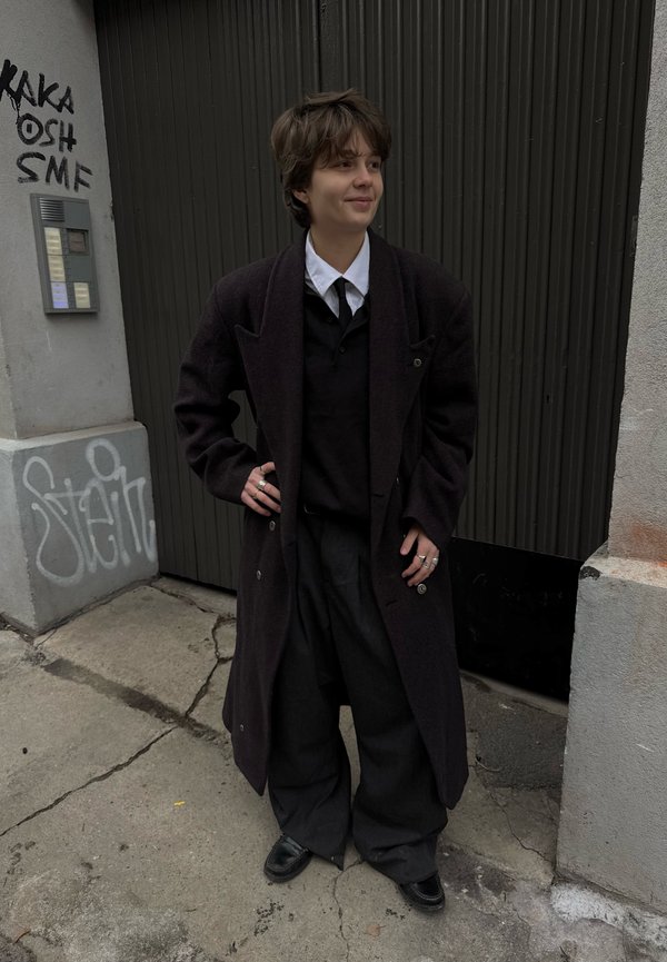 Persoon staand buiten bij een met graffiti bedekte muur, gekleed in een donker overjas, wit overhemd, zwarte stropdas, losse broek en meerdere ringen, handen in de zij.