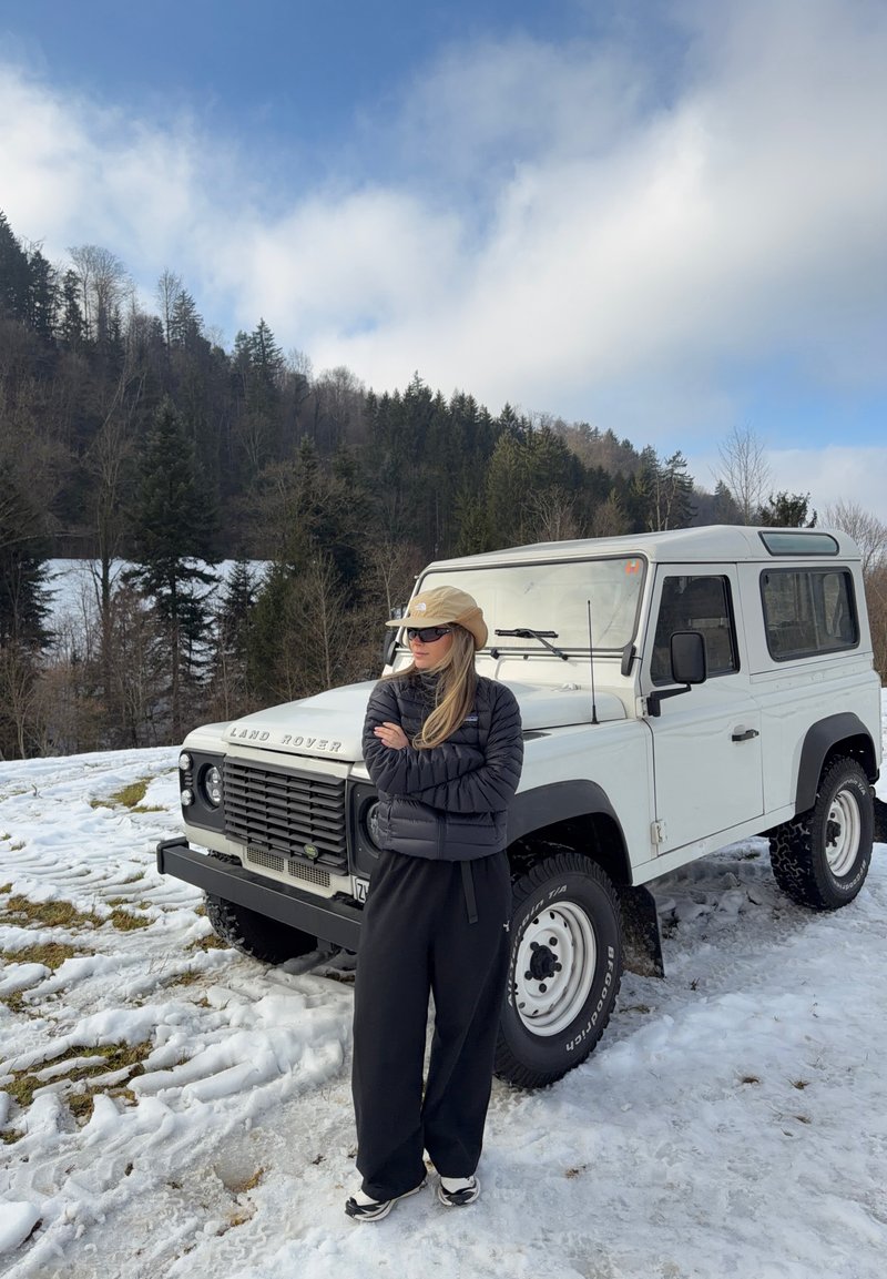 Femme en vêtements d'hiver et lunettes de soleil, les bras croisés, debout à côté d'un Land Rover blanc sur un sol enneigé près de collines boisées.