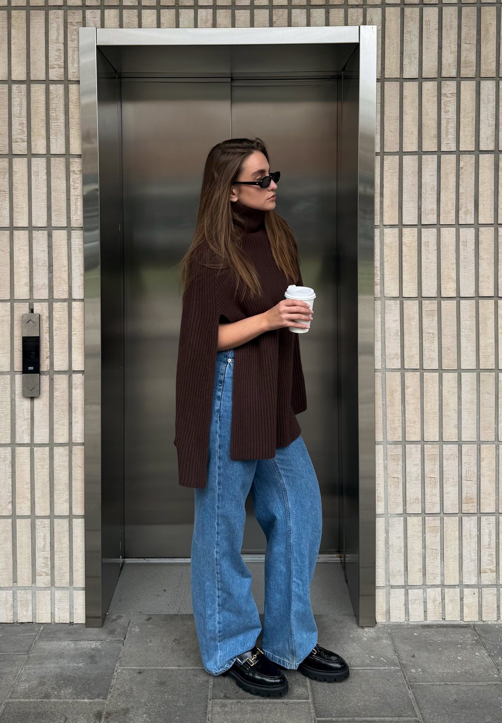 Barna bordázott pulóver magas gallérral, széles kék farmer, és fekete platformos cipő. Kávés bögrét tart az lift közelében.