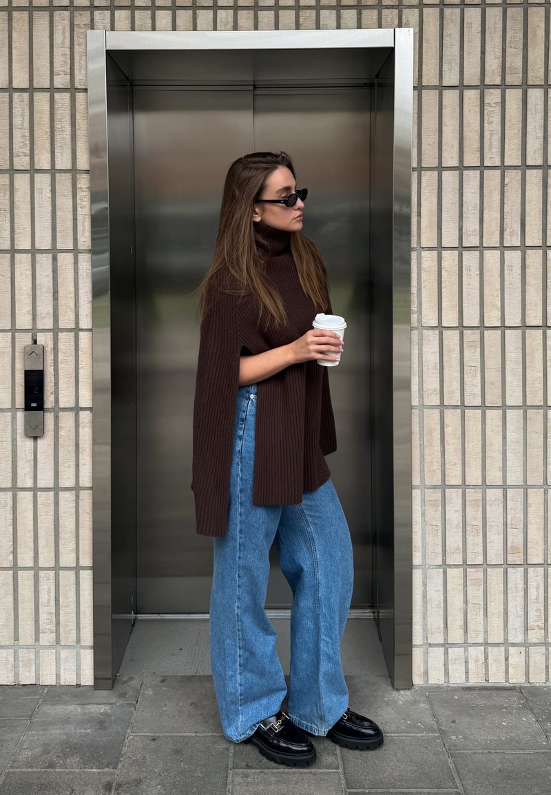 Pull marron côtelé à col montant, jean large bleu, et chaussures noires compensées. Tenant une tasse de café près d'un ascenseur.