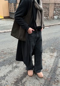 Manteau noir surdimensionné, écharpe grise, chemise noire, jean noir ample et chaussons beige. Le matériau est doux ; le design est décontracté et superposé.