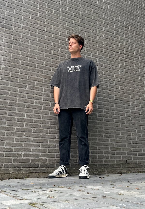 Camiseta negra de gran tamaño con texto blanco, vaqueros negros de corte recto y zapatillas deportivas con estampado, frente a una pared de ladrillos gris.