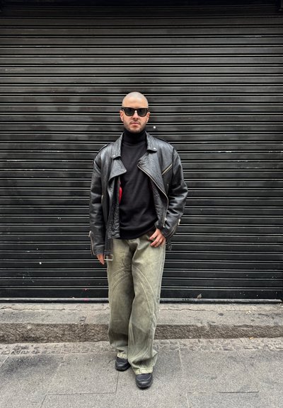 Chaqueta de motorista de cuero negro, cuello de tortuga negro, pantalones anchos oliva con detalles de costura, gafas de sol negras, de pie contra un fondo negro texturizado.