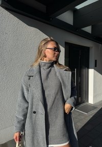 Palton gri din lână purtat peste o rochie cu guler înalt cu dungi alb-negru. Ochelari pătrați, groși și cercei mari cerc completează ținuta.