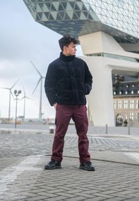 Black quilted jacket with a high collar, paired with maroon trousers and black trainers. The background features modern architecture and wind turbines.