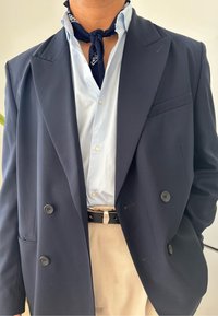 Blazer azul marinho de dupla face com botões pretos contrastantes, camisa listrada azul clara e lenço de seda escura com padrão sobre calças bege.