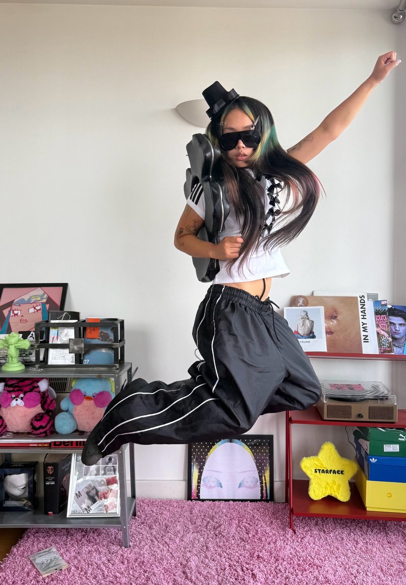 Person wearing sunglasses, black hat, and black trousers jumps in a room with pink carpet, shelves holding books, toys, and artwork.