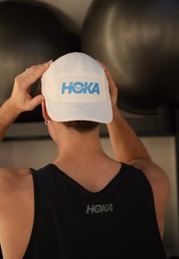 Casquette blanche avec logo « HOKA » bleu, présentant un design structuré et une texture lisse, portée par une personne portant un débardeur noir.