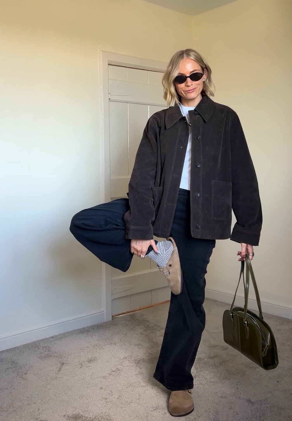 Jachetă din suede maro, cămașă albă, pantaloni negri evazați, papuci bej cu șosete cu dungi, poșetă verde, păr scurt și ondulat, și ochelari de soare.