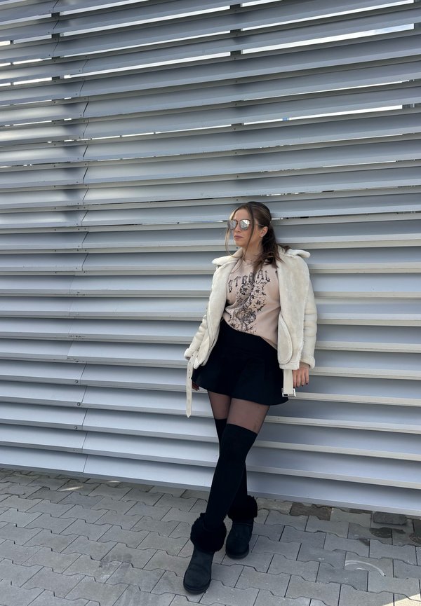 Chaqueta de piel sintética blanca sobre un sudadera gráfica beige, falda corta negra y calcetas negras hasta el muslo con botas negras hasta el tobillo. Gafas de sol plateadas.