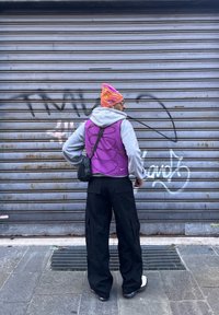 Personne debout de dos, portant un bonnet rayé orange, un gilet violet sur un sweat à capuche gris, un pantalon cargo noir et des baskets blanches, près d'un volet métallique.