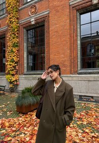 Brun dobbelspent frakk med store slag, brukt over en lys skjorte. Omgitt av høstløv, murvegg og store vinduer.