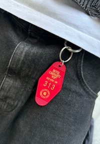 Llavero de hotel rojo con texto en relieve "HOTEL CARHARTT WIP" y el número "313", atado a un anillo de metal en unos jeans oscuros.