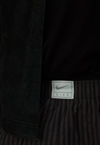 Chaqueta de ante negra con costuras visibles y una capa negra texturizada por debajo; etiqueta gris de Nike en pantalones cortos de rayas negras.