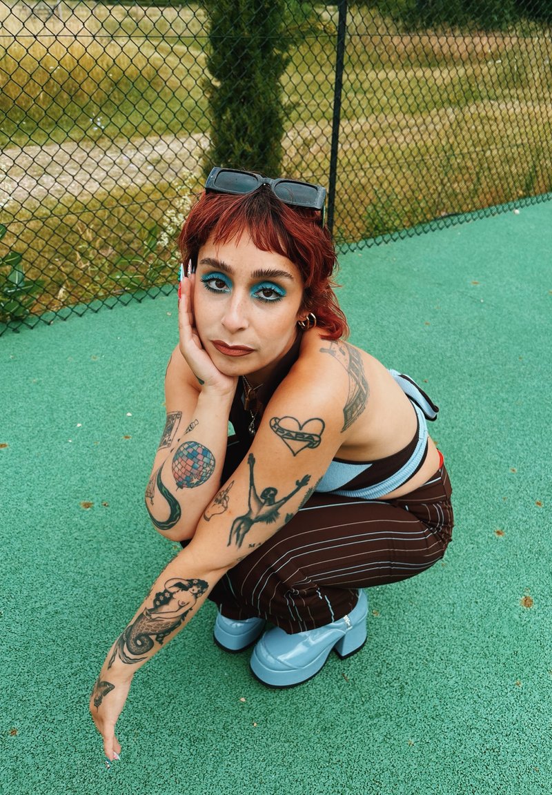 Personne aux cheveux roux et au maquillage des yeux bleu accroupie sur un terrain vert portant un pantalon rayé, des chaussures bleu clair et des tatouages sur les deux bras.