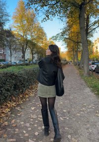 Žena oblečená v čiernej koženej bunde, sivej denimovej sukni, čiernych pančuchách a čiernych čižmách po kolená, kráčajúca po ceste lemovanej stromami s jesenným lístím.