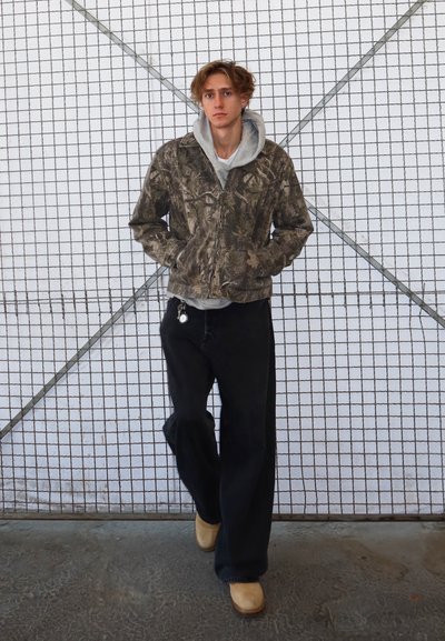 Chaqueta de camuflaje con una sudadera gris por debajo, pantalones anchos negros y zapatos beige tipo slip-on, todo contra un fondo de patrón de cuadrícula.