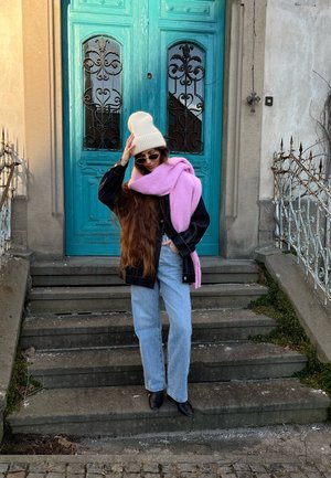 Chaqueta negra, vaqueros azul claro, bufanda rosa y gorro blanco. Cabello largo y castaño que cae en cascada. De pie en escalones de piedra, puerta turquesa de fondo.